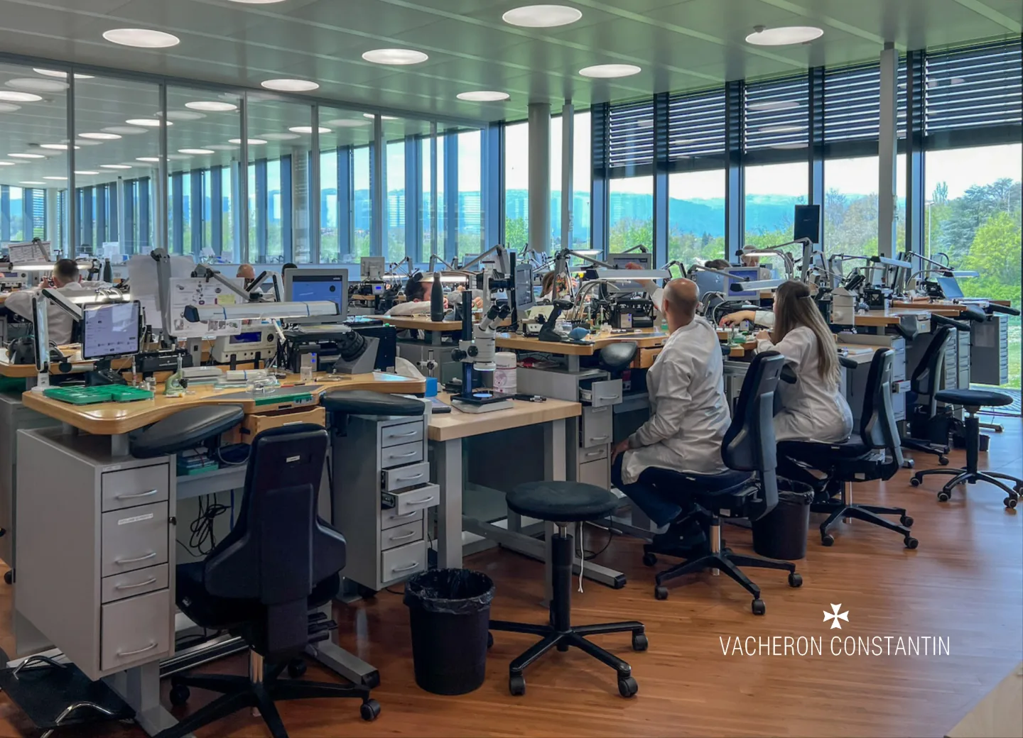 A Vacheron Constantin workshop: rows of watchmakers' benches with microscopes and task lamps, along a wall of windows.