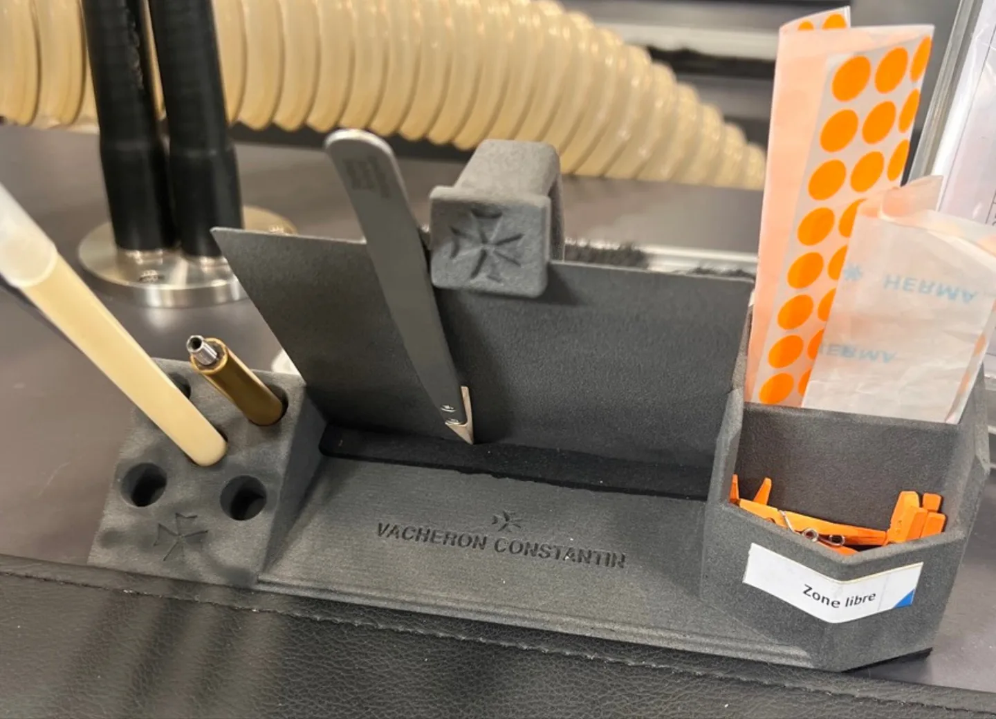 A grey 3D-printed bench stand holding tweezers and blades, engraved with the Vacheron Constantin name and Maltese cross.